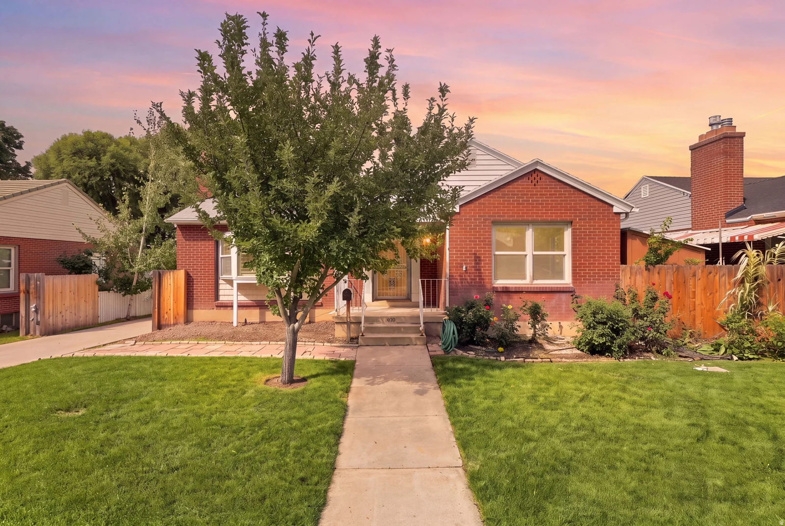 Red brick exterior with white trim, mature tree, concrete walkway, green lawn, and wood fencing